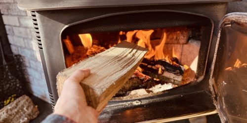 Hand adding a log to a burning fire inside a wood stove, providing warmth and comfort in a cozy indoor setting.